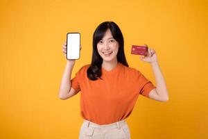dinámica asiático mujer en su 30, revelador teléfono inteligente y crédito tarjeta en amarillo antecedentes. eficiente en línea pago compras. foto