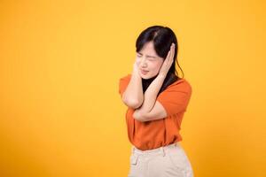 joven asiático mujer vistiendo naranja camisa cerrado oído con mano para silencio a oír un ruido sonido en contra amarillo antecedentes. infeliz desesperado gesto concepto. foto