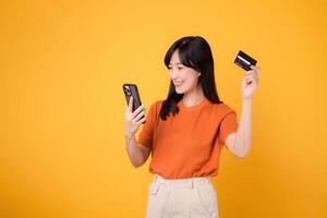 Excited Asian woman 30s in orange shirt, using smartphone and holding credit card on yellow background. Fast online shopping. photo