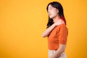 Vigilant young Asian woman in her 30s, wearing an orange shirt, holds her pain shoulder on yellow background. Neck ache therapy medical office syndrome concept. photo