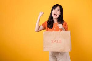 Join the celebration of love and incredible deals for joyful shopping experience. Trendy asian woman reveals her purchases, embodying the excitement of smart shopping. isolated on yellow background. photo