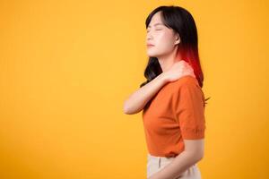 Aware young Asian woman in her 30s, wearing an orange shirt, holds her painful neck on yellow background. Neck ache therapy medical office syndrome concept. photo