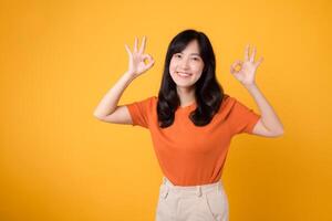 A young Asian woman in her 30s, wearing an orange shirt, showcases the okay sign gesture on a sunny yellow background. Hands gesture concept. photo