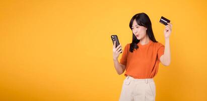Joyful Asian woman 30s in orange shirt, using smartphone and holding credit card on yellow background. Seamless online payment. photo