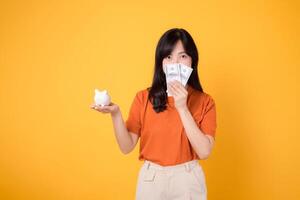 Joyful Asian woman in her 30s kisses piggy bank, holds cash money dollars on vibrant yellow background. Wealth saving concept. photo