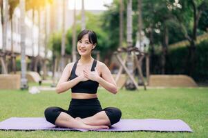 Portrait fit beautiful young asian woman 30s wearing black sportswear holding hands on chest while listening relax music and sitting on yoga mat in public park. Healthcare wellness concept. photo