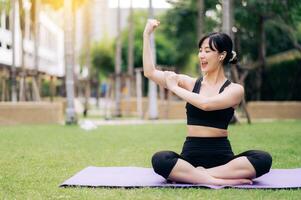 Portrait fit beautiful young asian woman 30s wearing black sportswear listening relax music and showing strong health gesture at her arm while do yoga on yoga mat in the outdoor park. photo