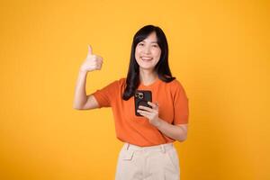 energético joven asiático mujer en su 30, vistiendo naranja camisa, utilizando teléfono inteligente con pulgar arriba mano firmar en vibrante amarillo antecedentes. nuevo móvil aplicación concepto. foto