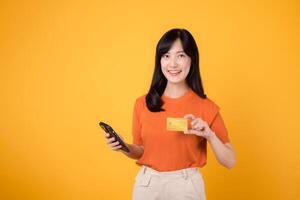 animado asiático mujer en su 30, empleando teléfono inteligente y exhibiendo crédito tarjeta en vibrante amarillo antecedentes. acelerado en línea compras pago proceso. foto