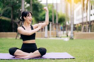 Portrait fit pretty young asian woman 30s wearing black sportswear listening relax music and showing strong health gesture at her arm while do yoga on yoga mat in the outdoor park. photo