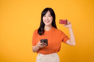 emocionado asiático mujer 30s en naranja camisa, utilizando teléfono inteligente y demostración crédito tarjeta en amarillo antecedentes. rápido en línea compras. foto