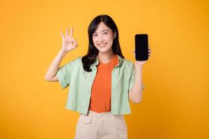 el futuro de nuevo móvil solicitud con joven asiático mujer en su 30, vistiendo naranja camisa y verde saltador, revelador teléfono inteligente pantalla con un bueno mano gesto en amarillo estudio antecedentes. foto