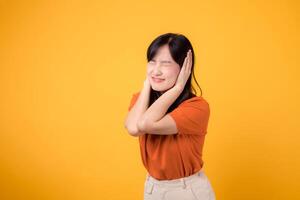 Stressed young asian woman in pain, holding her ear due to loud noise. Unhappy expression on isolated yellow background. photo