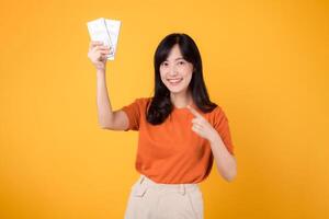 Dynamic Asian woman in her 30s points to cash money dollars on vibrant yellow backdrop. Wealth and prosperity concept. photo