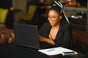 joven niña en lentes Asombrosamente mirando en ordenador portátil a cafetería. africano americano niña sentado en restaurante con ordenador portátil y taza en mesa. retrato de sorprendido dama con oscuro Rizado pelo en auriculares foto