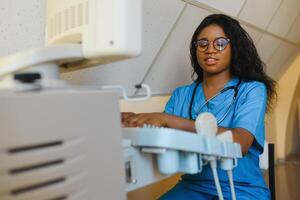 Young cheerful African woman operator of an ultrasound scanning machine analyzing diagnostics results of patient. Young smiling African doctor working on a modern ultrasound equipment. photo