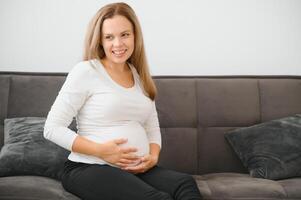 hermosa embarazada mujer sentado a sofá y acuerdo mano en barriga. foto