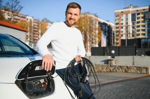 young handsome man holding charging cable at electric charging station point standing near his new car. photo