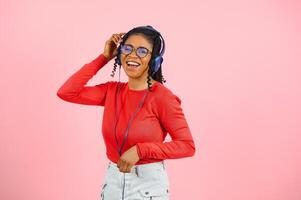 Pretty african american young woman with bright smile dressed in casual clothes, glasses and headphones dance over pink background photo
