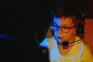 Child online broadcasts computer game, boy streams in headphones on rgb lighting background photo