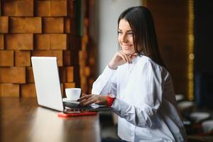 joven mujer exitoso gerente es hablando vía móvil teléfono con posible financiero empresa empleado y leyendo su currículum en portátil ordenador portátil computadora durante remoto entrevista en trabajo colaborativo espacio foto