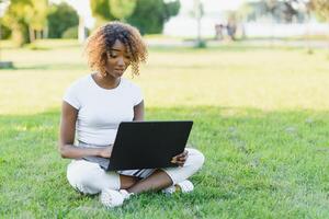 pensativo linda mezclado hembra internacional estudiante con Rizado pelo es sentado en Fresco césped con moderno ordenador portátil en público parque, propensión en manzana árbol y con nostalgia mirando aparte durante su descanso foto