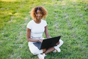 Thoughtful cute mixed female international student with curly hair is sitting on fresh grass with modern laptop in public park, leaning on apple tree and wistfully looking aside during her break photo