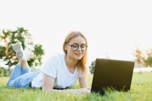 Beautiful and happy young woman lying on the grass surrounded by technology devices photo