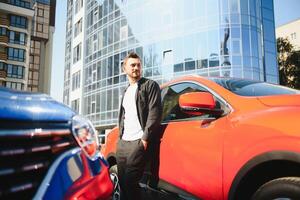 An attractive man with a beard stands next to his car in the parking lot. He is thoughtful, and looks away, someone is waiting. Summer day photo