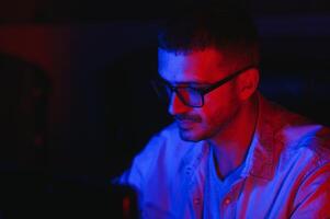 Pro Gamer. Millennial Guy Using Desktop Computer Playing Online Videogame Sitting At Night At Home. Neon Lights, Selective Focus photo