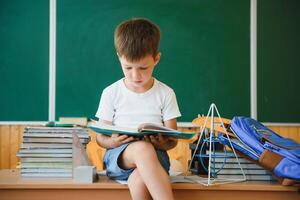 contento linda inteligente chico es sentado a un escritorio. niño es Listo a responder con un pizarra en un antecedentes. Listo para escuela. espalda a escuela. libros en escritorio foto