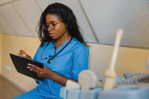 Young african-american female doctor in white coat using ultra ultrasound scanning machine and looking on the screen. African woman doctor working on modern ultrasound equipment. photo