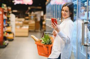 mujer haciendo tienda de comestibles compras a el supermercado, ella es propensión en el compras carro y conectando con su teléfono, aplicaciones y Al por menor concepto foto