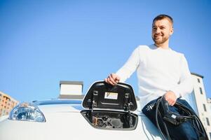 joven hermoso hombre participación cargando cable a eléctrico cargando estación punto en pie cerca su nuevo coche. foto