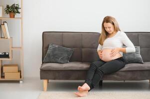 hermosa embarazada mujer sentado a sofá y acuerdo mano en barriga. foto