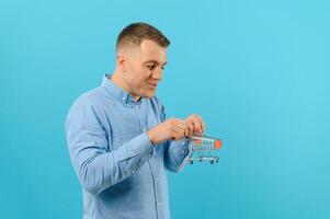 Young smiling man holding a mini shopping cart on a blue background. Sales concept. Place for text. photo