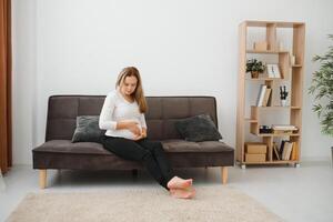 Beautiful pregnant woman sitting at sofa and keeping hand on belly. photo