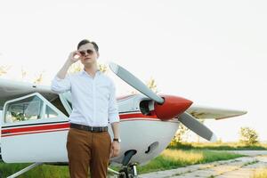 retrato de hermoso comercial piloto en capitán blanco uniforme en pie y mirando inteligente cerca pequeño privado helicóptero en un aterrizaje punto con privado chorro terminal a internacional aeropuerto foto