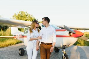Full-length image of beautiful young stylish couple near private plane. Walking on runway in airport in front of airplane. photo