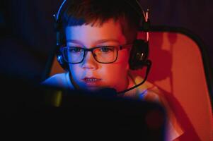 Child online broadcasts computer game, boy streams in headphones on rgb lighting background photo