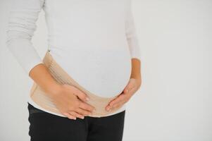 Close up of pregnant woman putting on a bandage at gray background with copy space. Orthopedic abdominal support belt concept. photo
