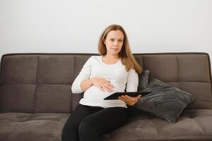 Joyful pregnant woman having fun with a tablet while sitting on sofa at home. photo