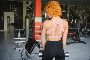 Close up photo. Beautiful sexy caucasian woman with perfect figure posing standing with her back near the window in the gym next to the rowing simulator and squats. photo