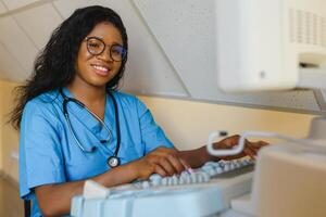 Young female African-american doctor working on modern ultrasound equipment. Operator of ultrasound scanning machine sitting and looking at the monitor, waiting for patient. photo