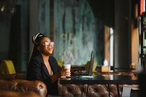 hermosa afro americano niña en casual ropa es utilizando un ordenador portátil y sonriente mientras sentado en café foto