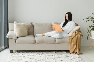 bonito joven mujer disfrutando leyendo un libro a hogar acostado en el sofá sonriente en Placer en casual ropa. foto
