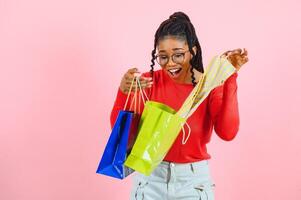 hermosa alegre africano americano mujer con pila de algo de papel pantalones en manos teniendo divertido disfrutando compras, aislado terminado pastel rosado antecedentes foto