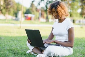 Young attractive dark-skinned college student wearing tank top and jeans sitting on the lawn at campus on sunny day, working on her thesis using laptop computer, looking busy and concentrated photo