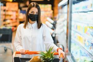 joven mujer con cara máscara caminando mediante tienda de comestibles Tienda durante covid-19 pandemia. foto