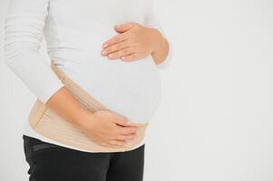 Close up of pregnant woman putting on a bandage at gray background with copy space. Orthopedic abdominal support belt concept. photo